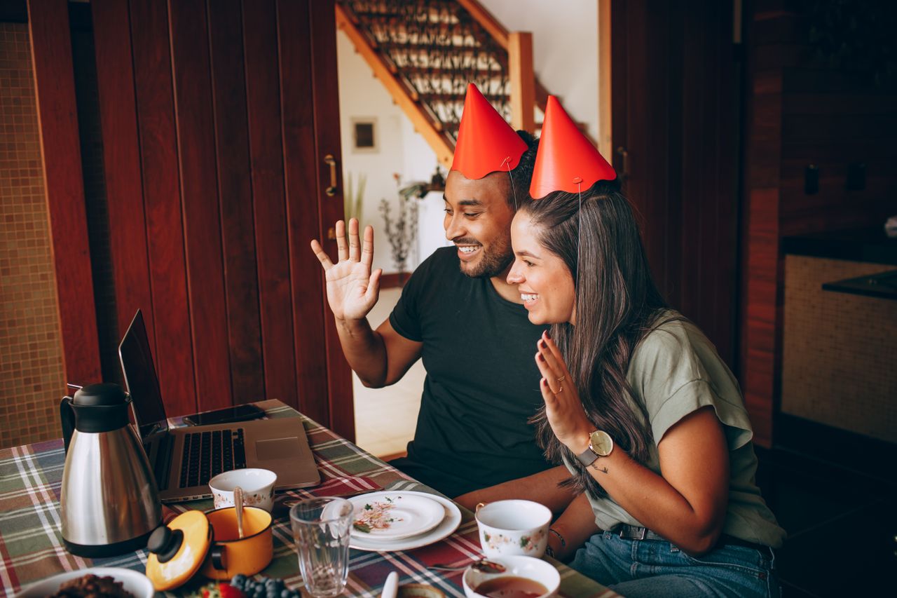 services-01 Two adults in a video call celebrating a birthday, wearing party hats and waving at a laptop indoors.